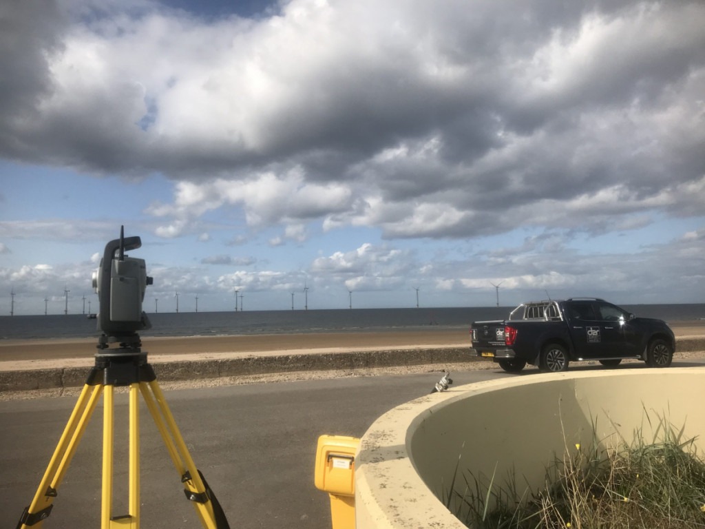 Total station on tripod with pick up truck on the sea fron with a wind farm in the distance