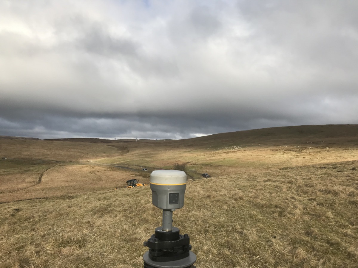 Grey GNSS receiver measuring a control point in the wilderness. There is a Yellow excavator near and wind turbines in the distance.