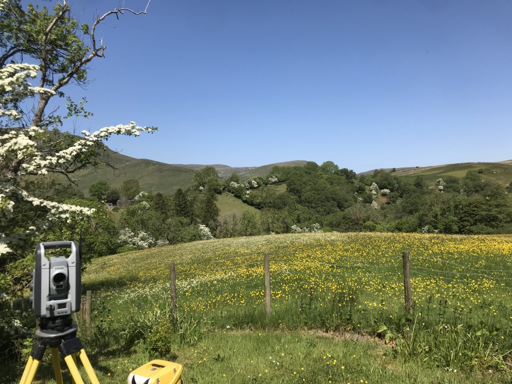 Grey total station on yellow tripod with rolling green hills in the background.