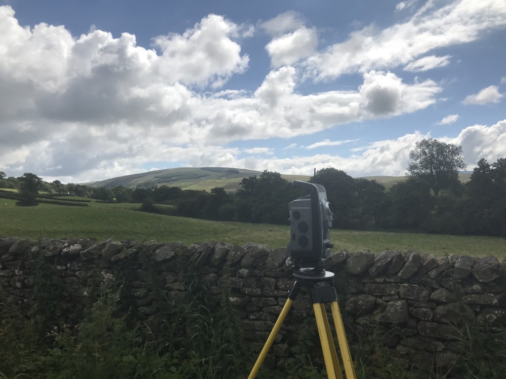 Grey Trimble S8 total station on yellow tripod in front of a dry stone wall with green fields and a treeline in the distance.