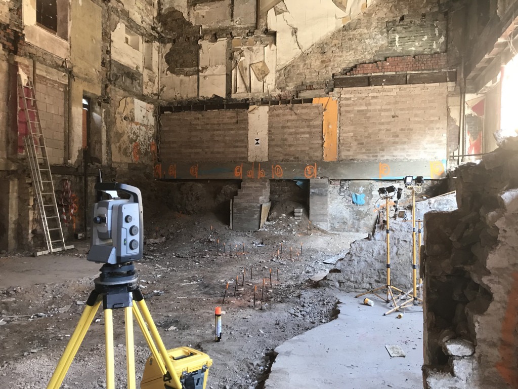 Grey S8 Total Station on Yellow tripod inside a derelict building basement. Orange painted Metal pins are inserted in the ground to mark the position of foundation piles to be installed.