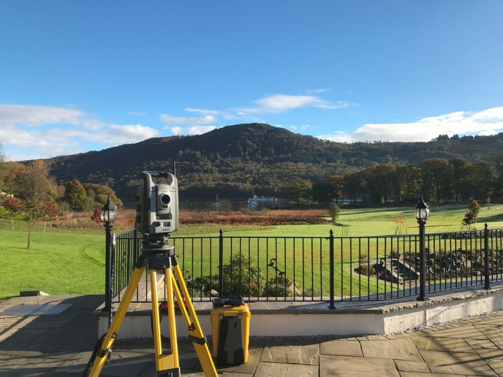 Total station on a yellow tripod in front of a garden railing with a rolling field down to a lake and mountain on the other side.