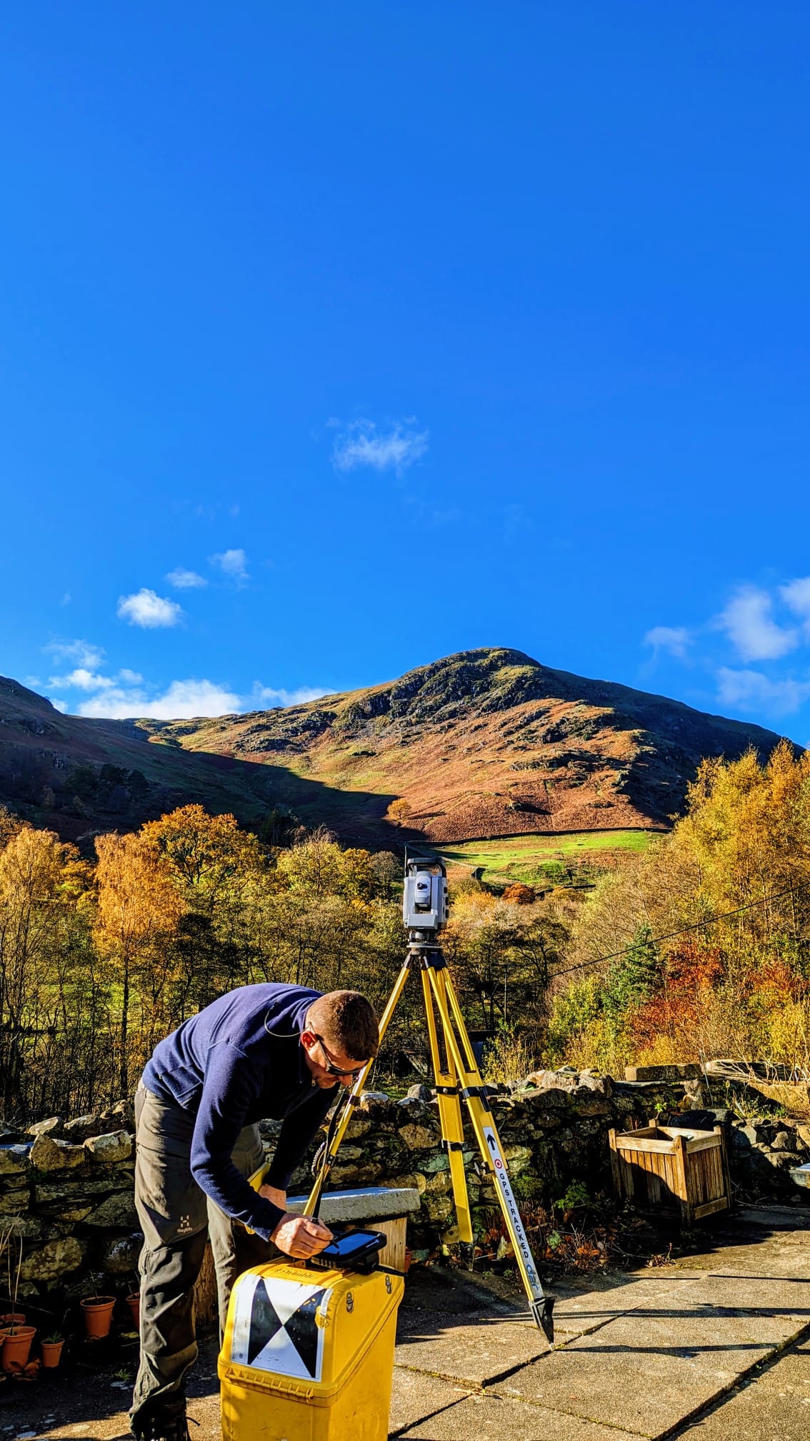 Surveyor working on his rugged handheld computer placed on a Yellow box on a patio. There is a total station on a Yellow tripod behind him. In the background is a mountain bathed in sunlight.