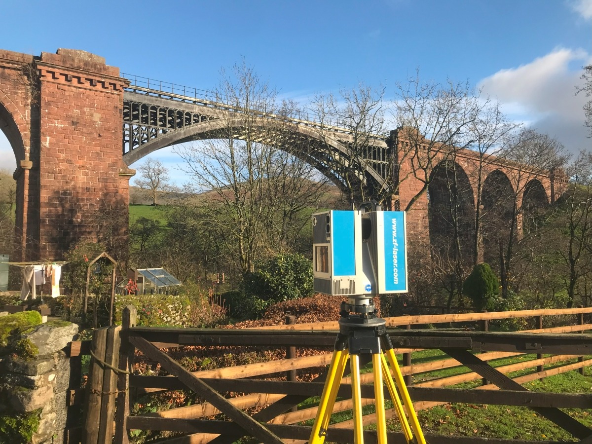 Blue 3D scanner on Yellow tripod in front of a wooden gate.  In the background is a sandstone railway viaduct with a black iron centre section.