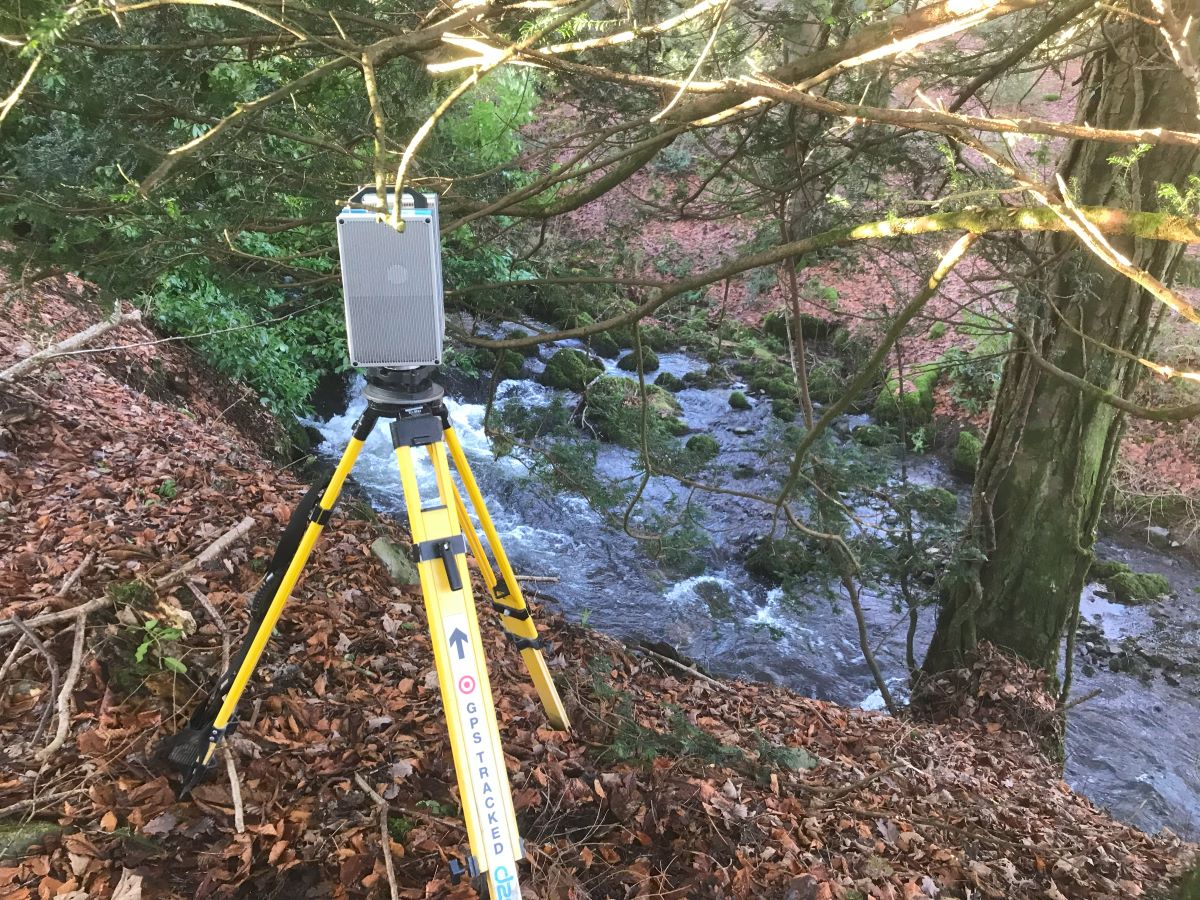 3D Scanner on a yellow tripod sited on an autumn leaf embankment overlooking a gushing stream with a pine tree hanging over.