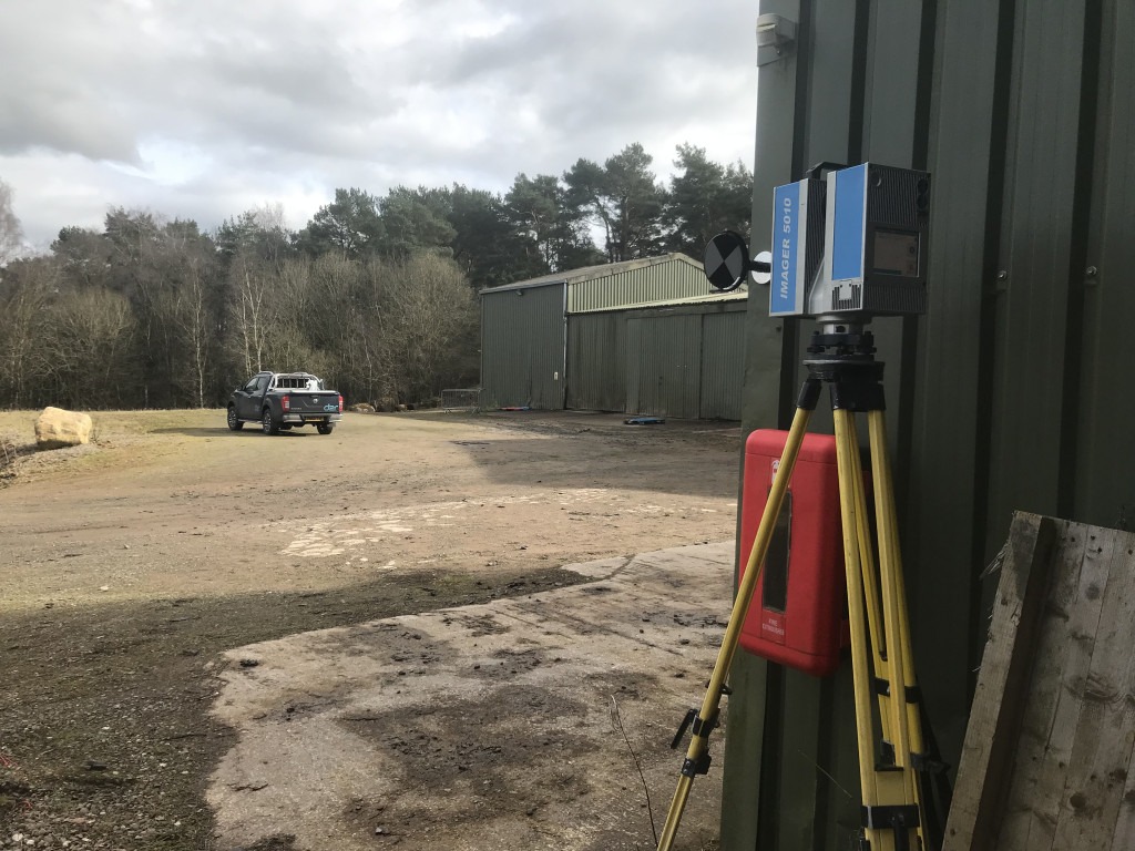 Green industrial sheds set in woodland. In the yard there is a blue pickup vehicle. Adjacent the nearest corner is a yellow tripod with a blue laser scanner operating. On the tin wall is a red fire equipment box.