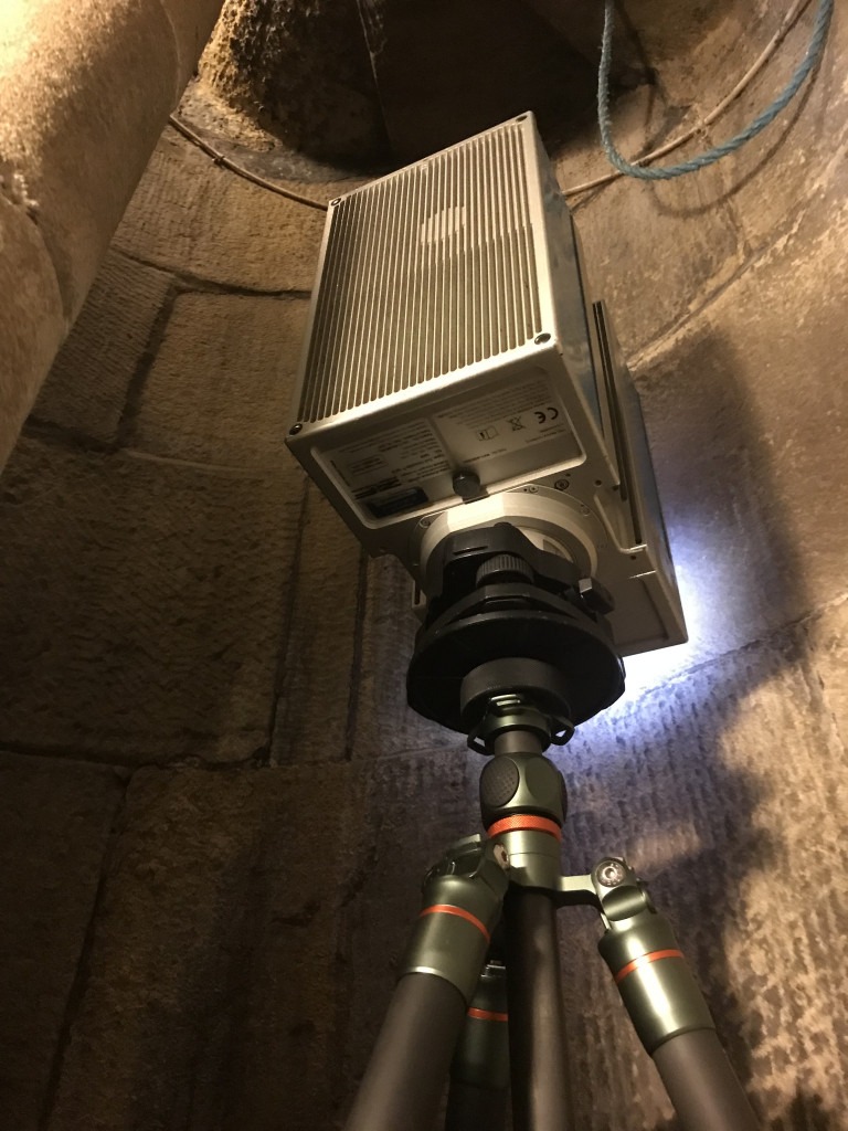 3D scanning surveying instrument on a tripod within a stone enclosed spiral staircase. There is a blue rope loop hanging over the scanner.