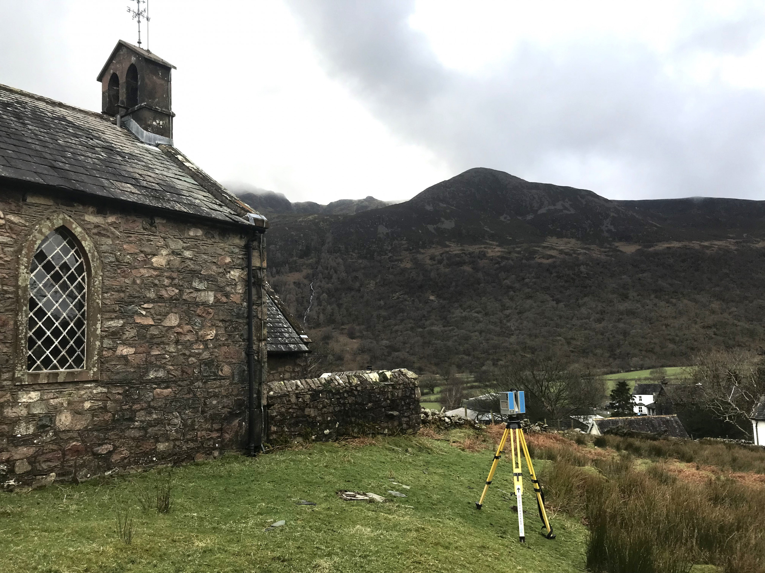 Blue 3D laser scanner on a yellow tripod measuring a stone church with mountains in the background under grey cloud.