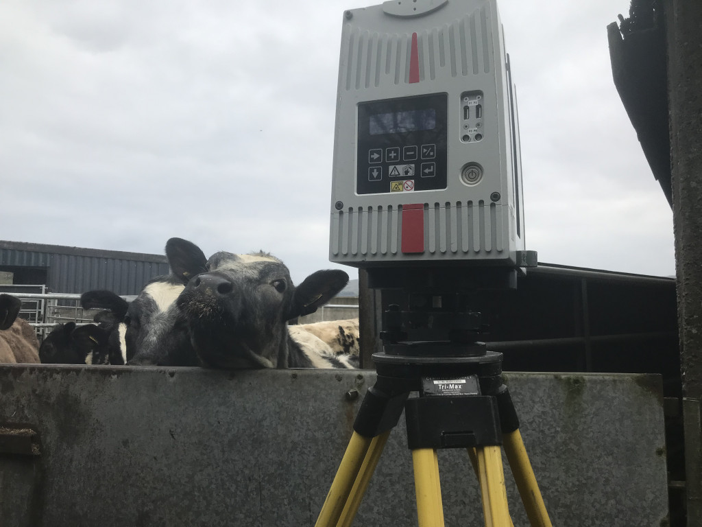 3D scanner on a yellow tripod in front of a concrete wall, on the other side but with its head over just out of licking reach, is a cow paying an interest to what's going on.