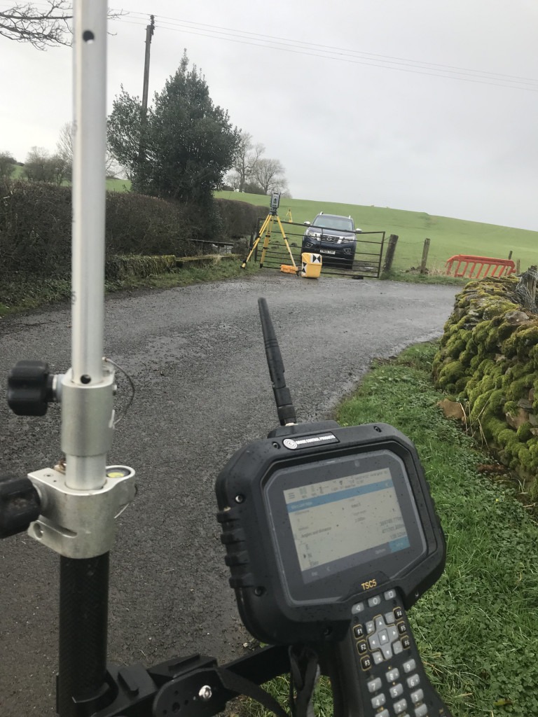 A survey pole with controller attached in the foreground, while the total station on a yellow tripod is on the other side of the road tracking the pole. Behind the total station is a gate and a pickup parked in a green field.